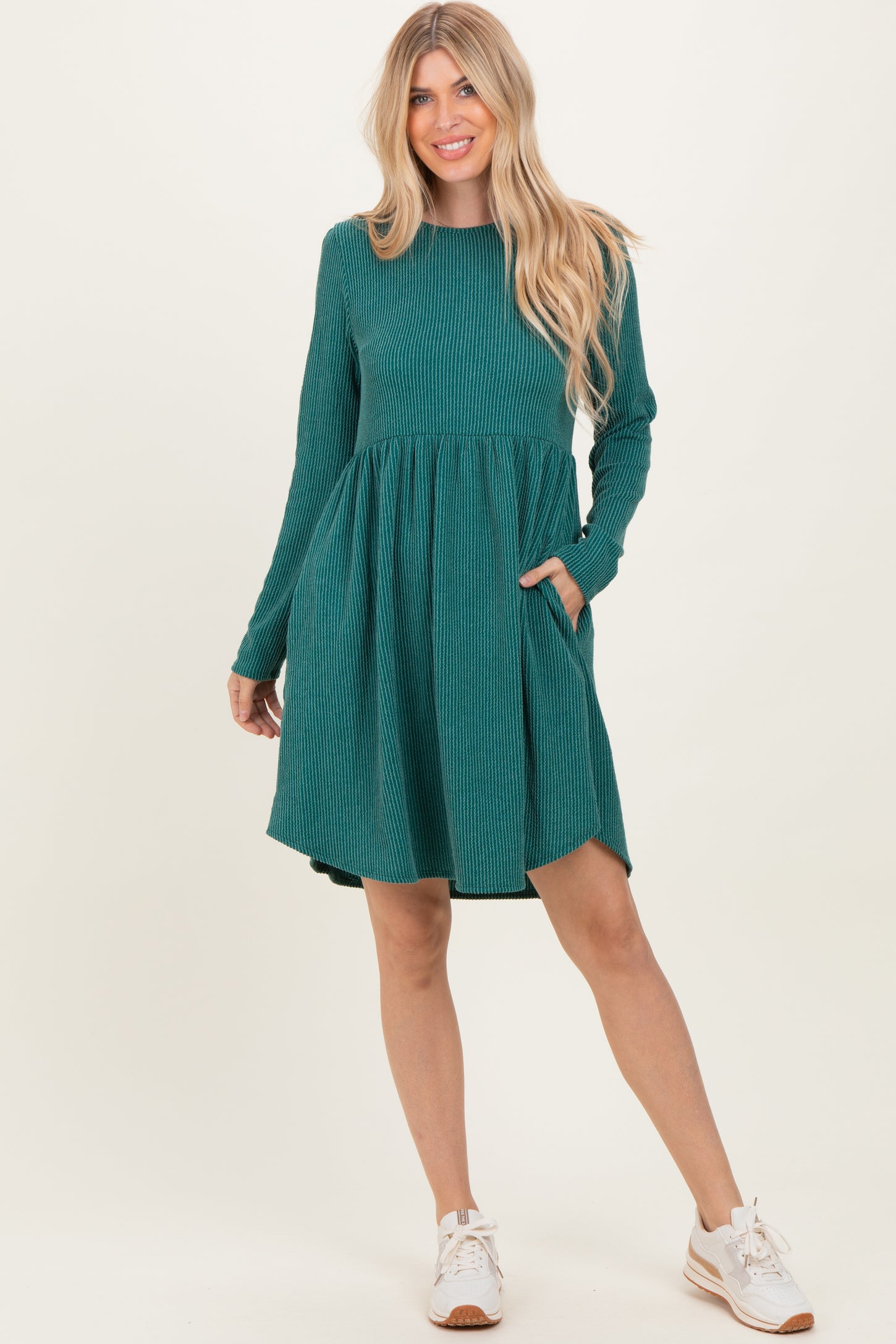 woman wearing a green ribbed long sleeve dress with a flared skirt and pockets, standing with one foot forward