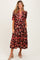 woman wearing a floral patterned brown maternity dress with a tiered skirt and red accents, posing with her hands in pockets