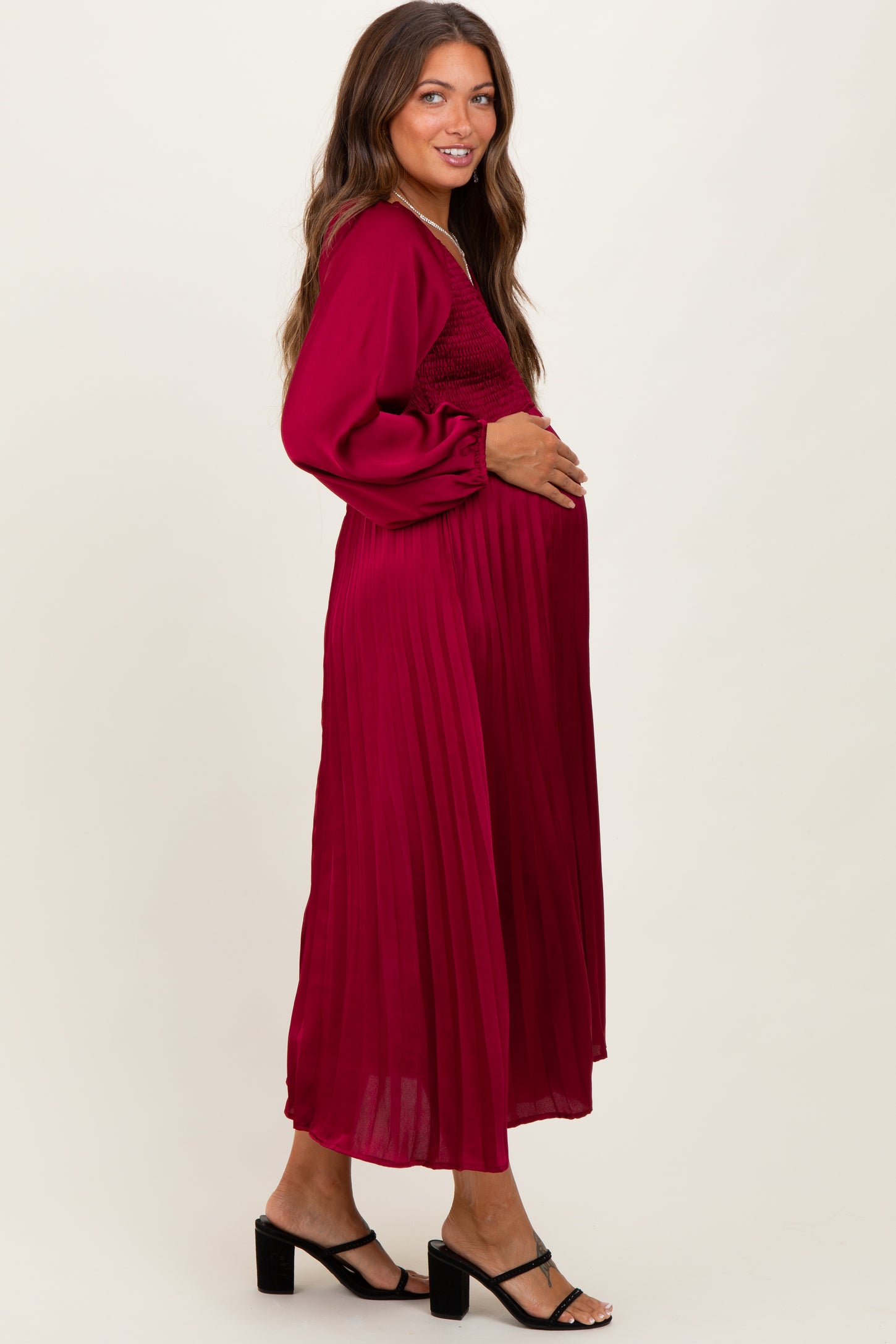 woman wearing a burgundy long sleeve maternity dress with pleats, gently cradling her baby bump