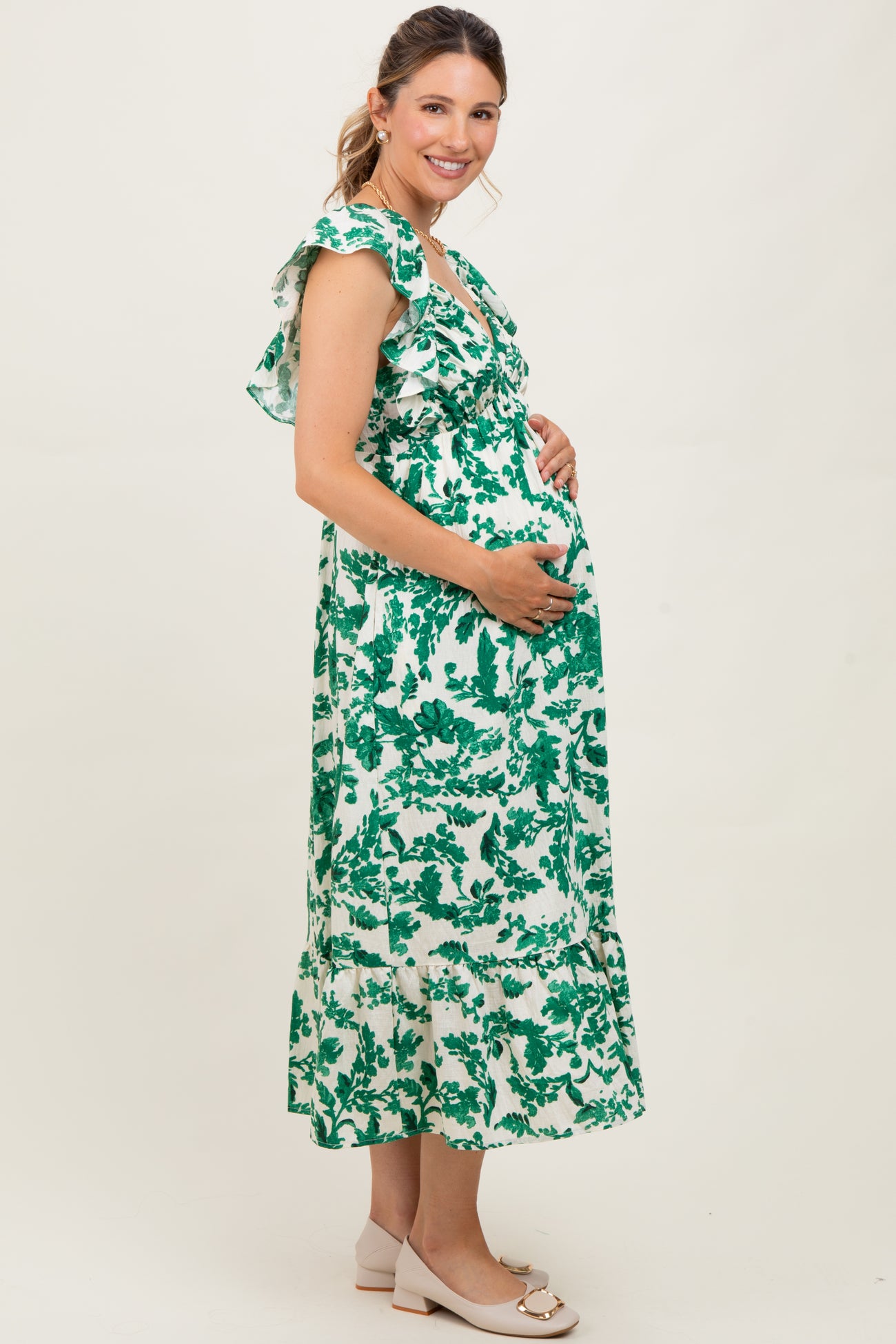 woman wearing a green and white floral maternity dress with ruffles, smiling and holding her baby bump