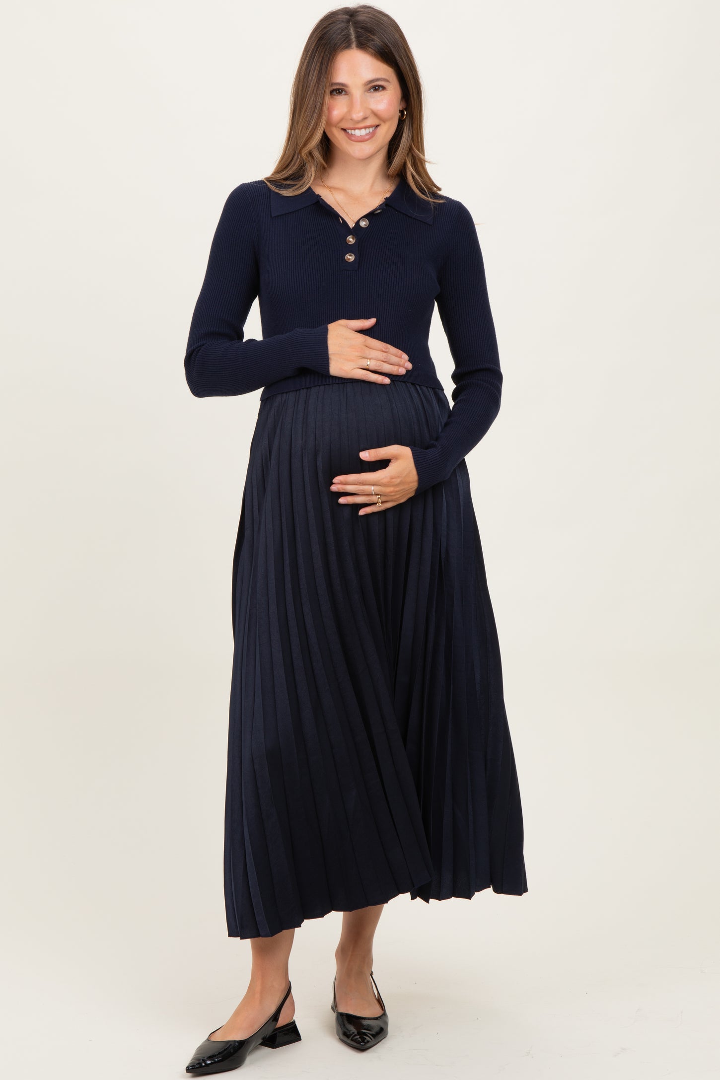 woman wearing a navy blue long sleeve maternity top with a collar and a pleated maxi skirt, holding her baby bump