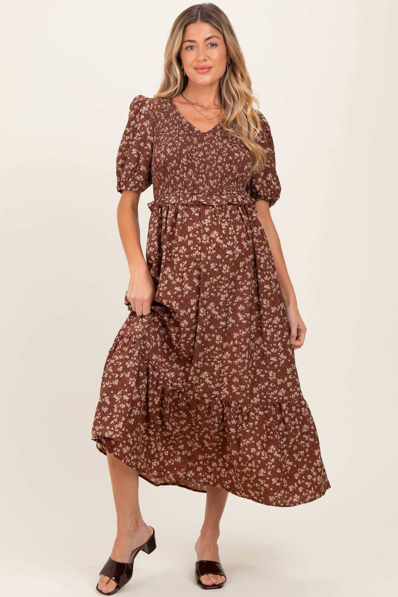 woman wearing a brown floral maternity dress with a v-neckline and puff sleeves, holding the skirt slightly