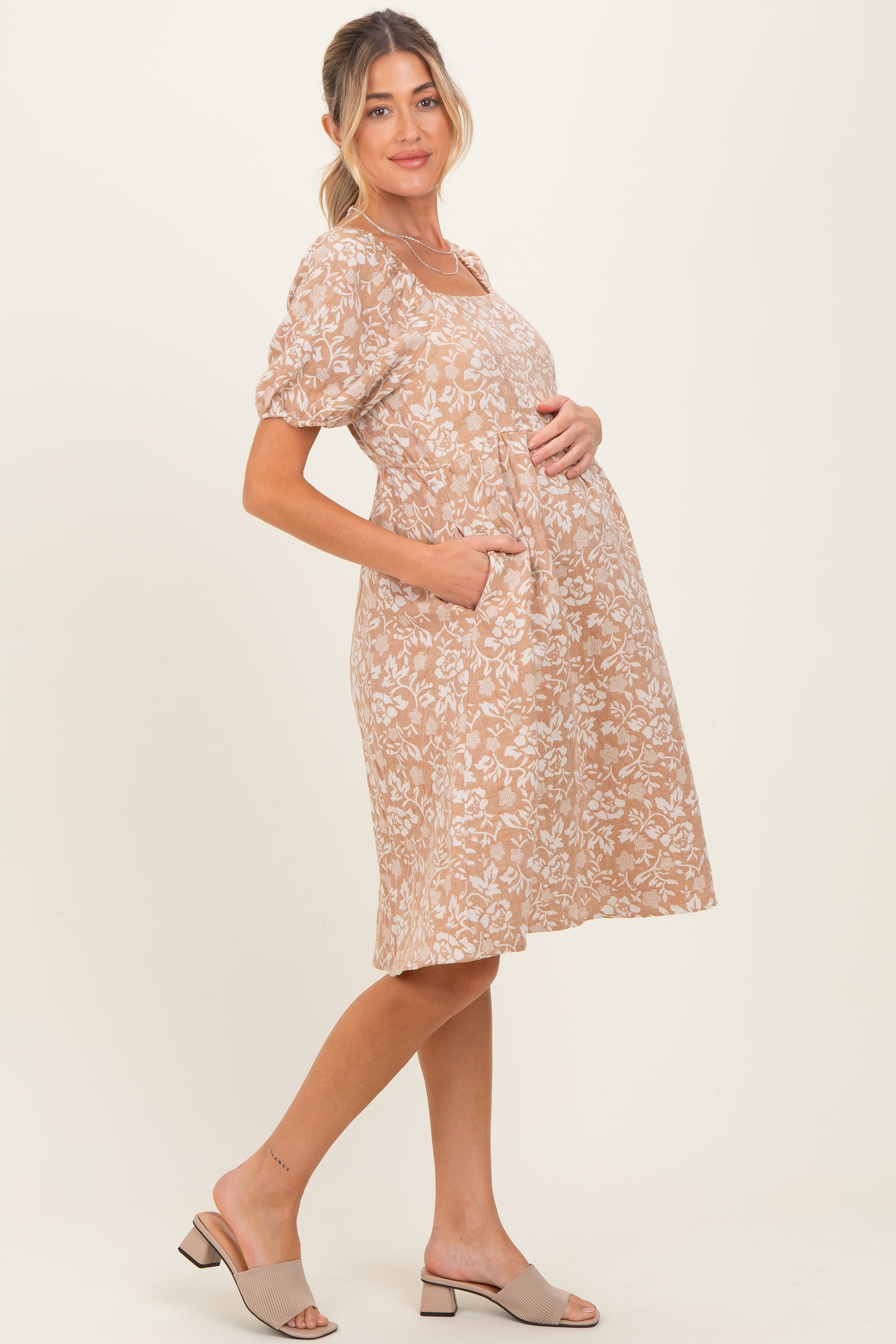 woman wearing a beige floral print maternity dress with short sleeves and pockets, showcasing her baby bump