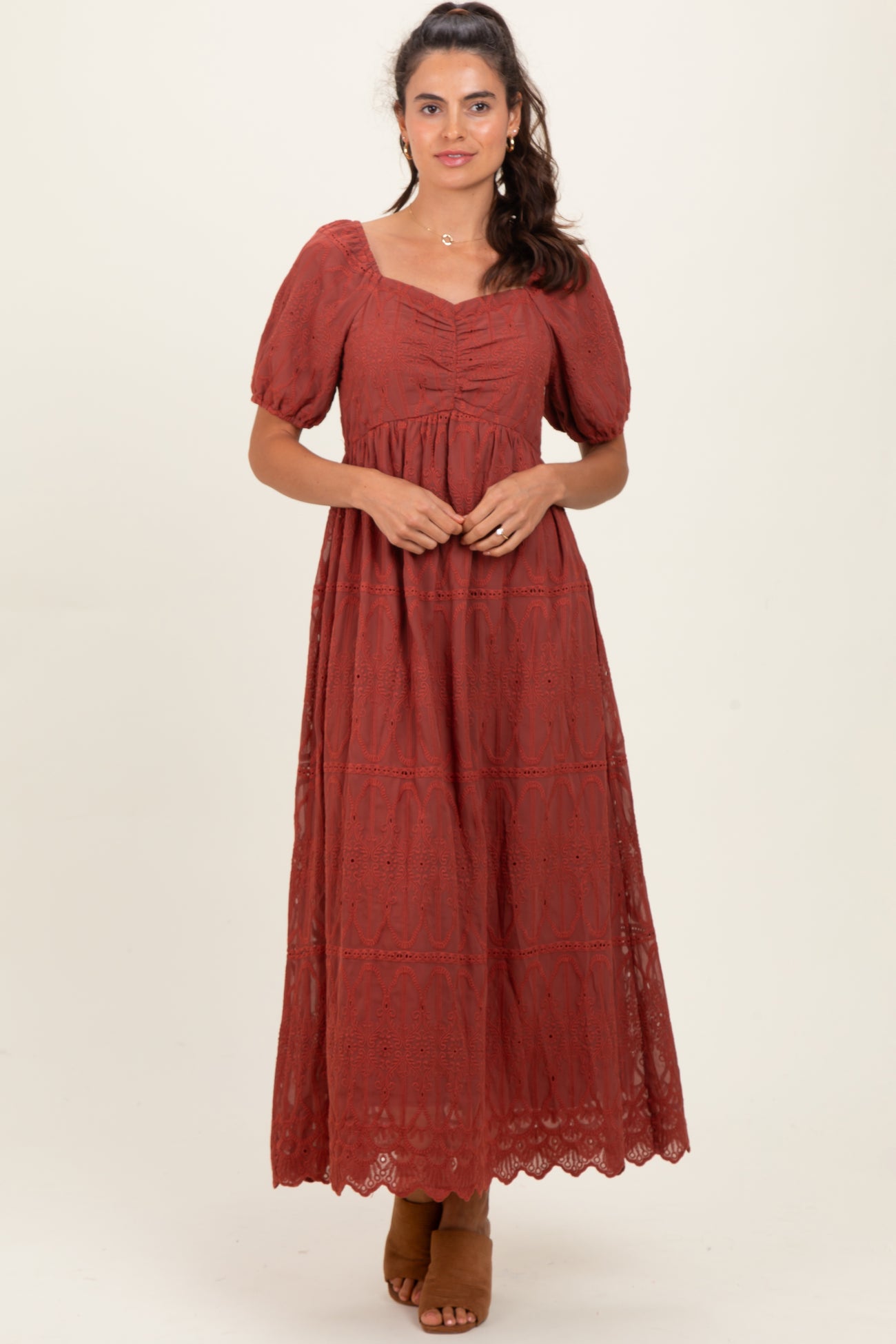 woman wearing a rust red maxi dress with puff sleeves and lace detailing, standing against a neutral background