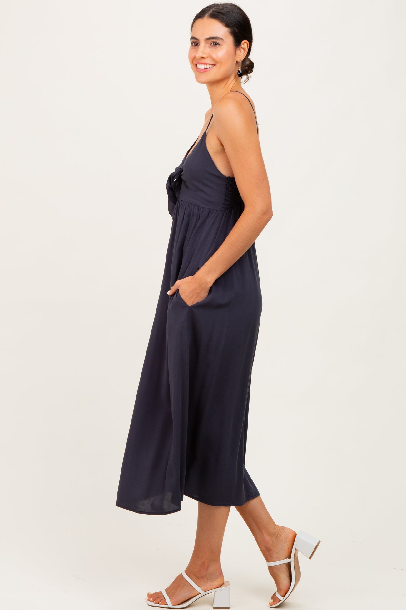woman wearing a navy blue sleeveless midi dress with a tie detail and pockets, standing sideways on a light background