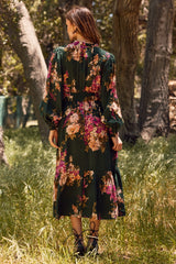 woman wearing a dark green floral maxi dress with long sleeves standing in a grassy area surrounded by trees