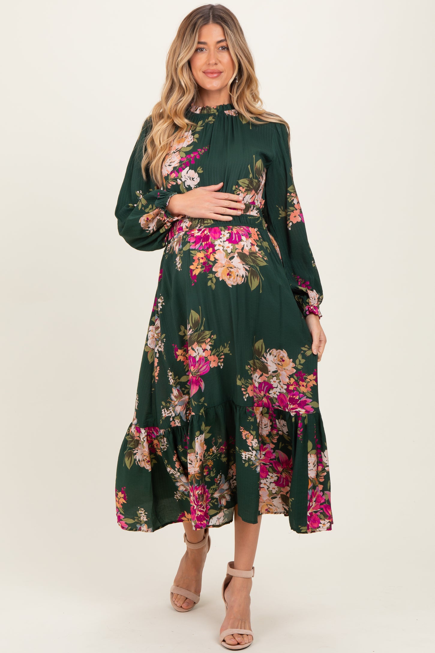 woman wearing a dark green floral maternity dress with long sleeves and a tiered skirt, gently cradling her baby bump