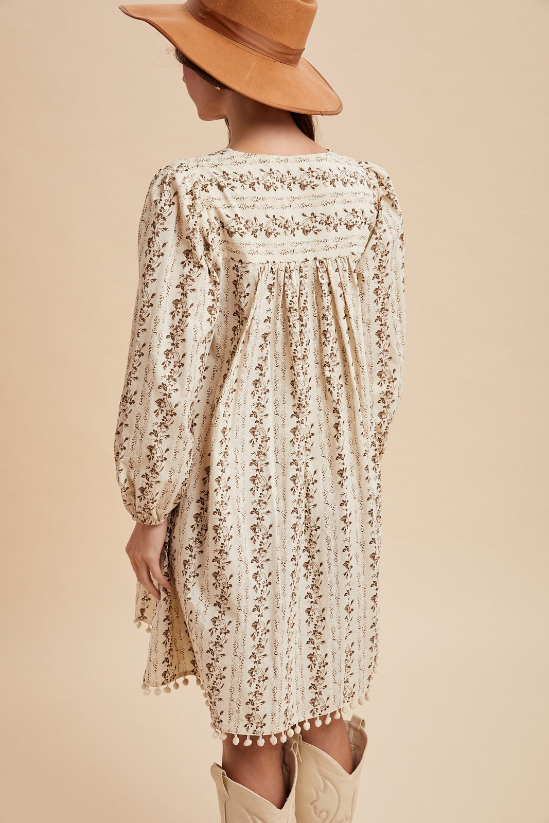 woman wearing a cream floral pattern long sleeve dress with a relaxed fit and pom-pom detail, styled with a brown hat