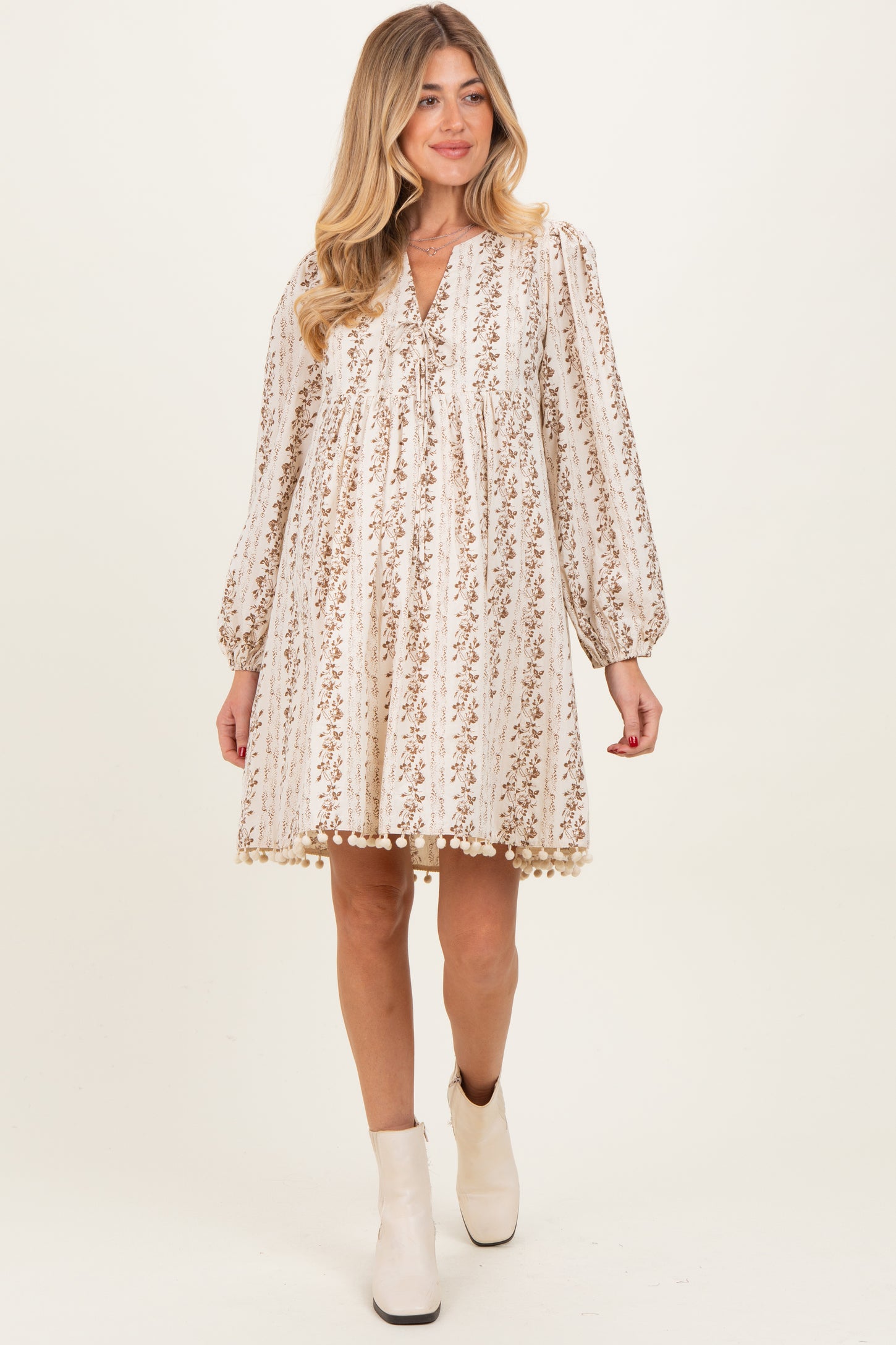 woman wearing a beige floral maternity dress with long sleeves and pom-pom trim, showcasing her baby bump