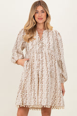 woman wearing a floral print maternity dress with long sleeves and pockets, featuring a gathered neckline and pom-pom trim