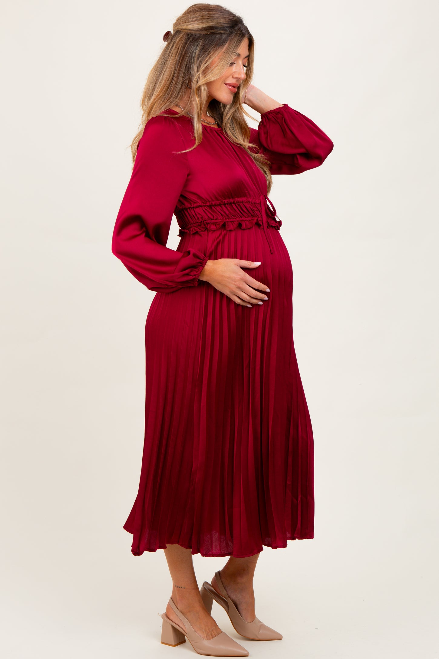 woman wearing a burgundy pleated maternity dress with long sleeves and a fitted waist, holding her baby bump