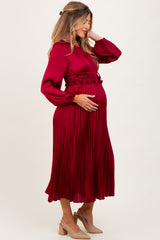 woman wearing a burgundy pleated maternity dress with long sleeves and a fitted waist, holding her baby bump