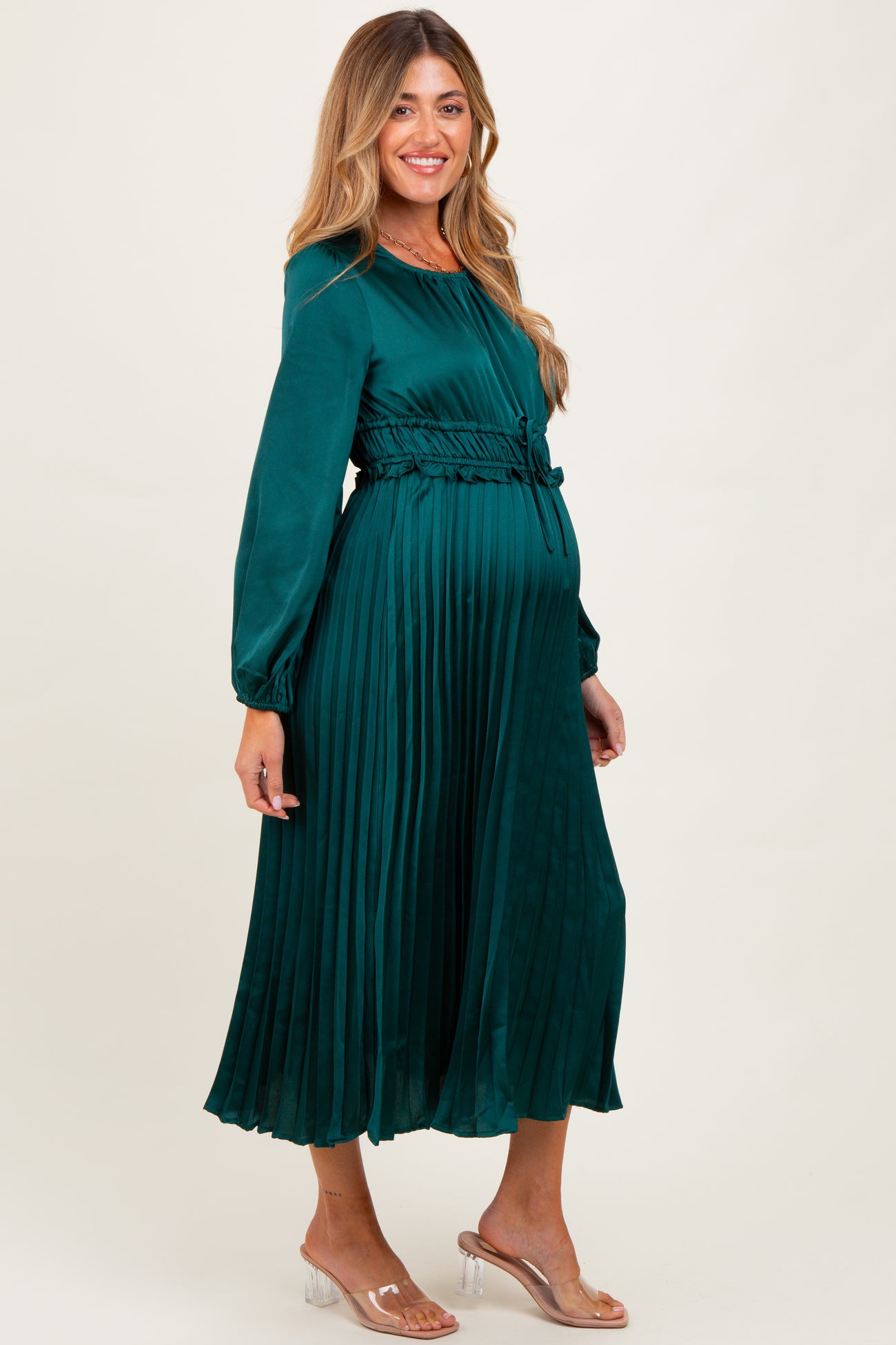 woman wearing a green pleated maternity dress with a tie waist and long sleeves, showcasing her baby bump