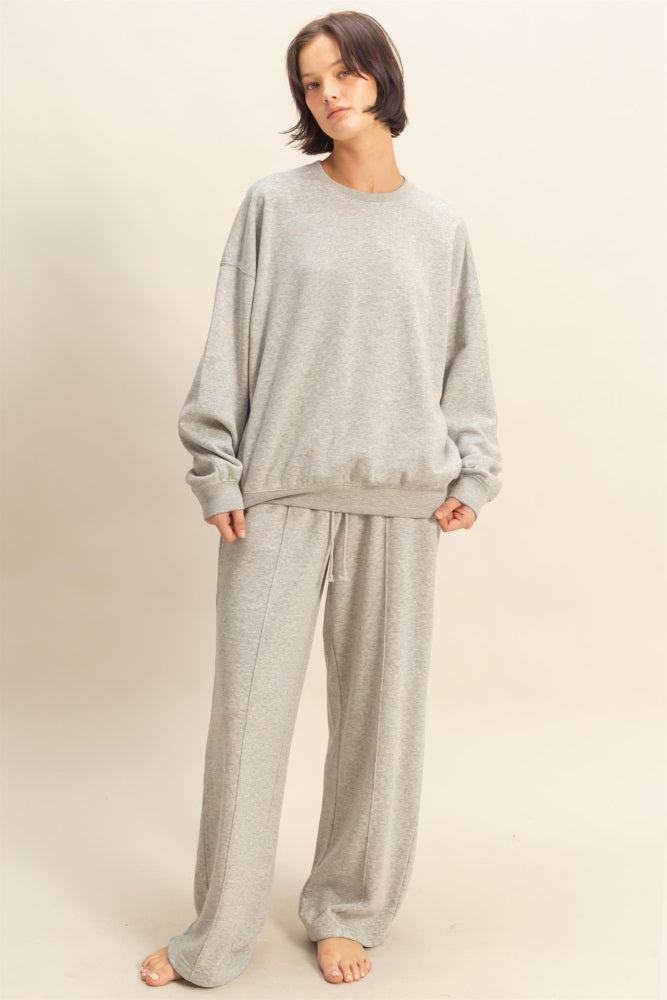 woman wearing a light grey oversized sweatshirt and matching sweatpants, standing barefoot against a neutral background