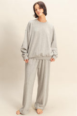 woman wearing a light grey oversized sweatshirt and matching sweatpants, standing barefoot against a neutral background