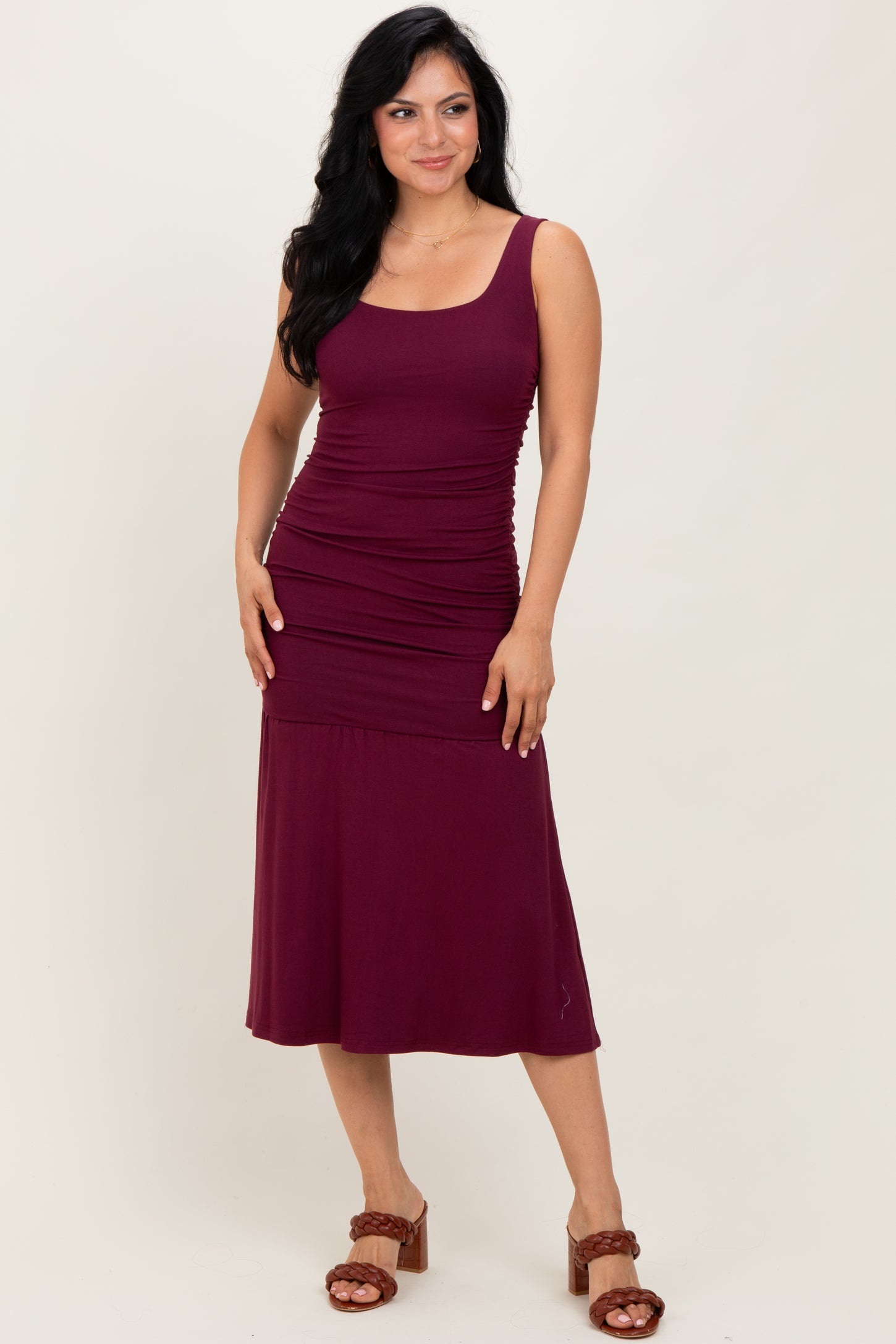 woman wearing a burgundy sleeveless midi dress with ruching details and standing with one hand on her hip