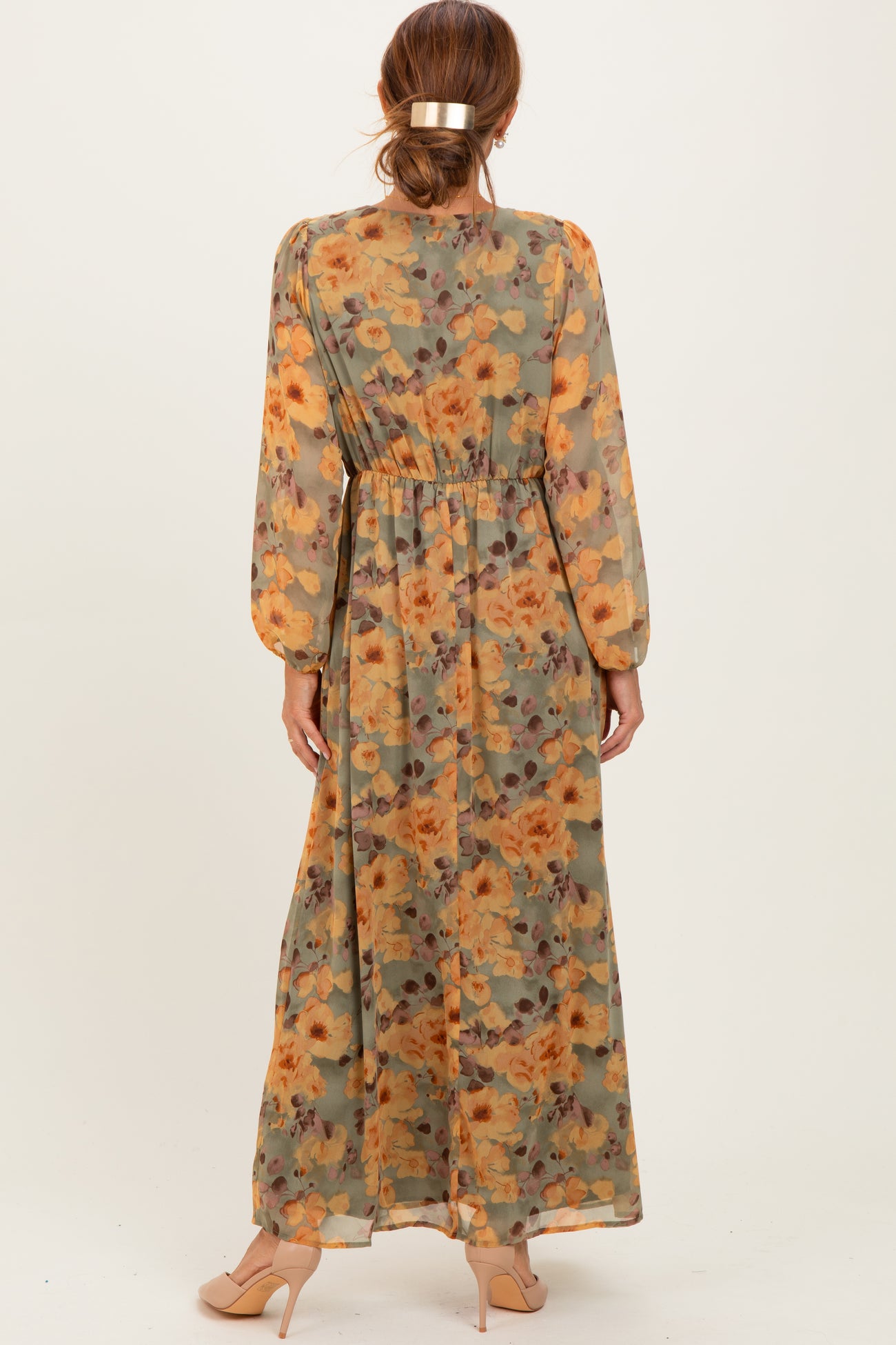 woman wearing a long floral maxi dress in shades of green and orange with long sleeves and a cinched waist from the back
