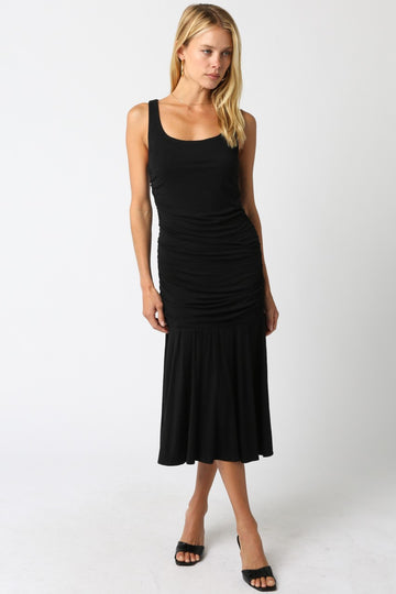 woman wearing a black fitted midi dress with ruching details and a flared hem, standing and looking at the camera