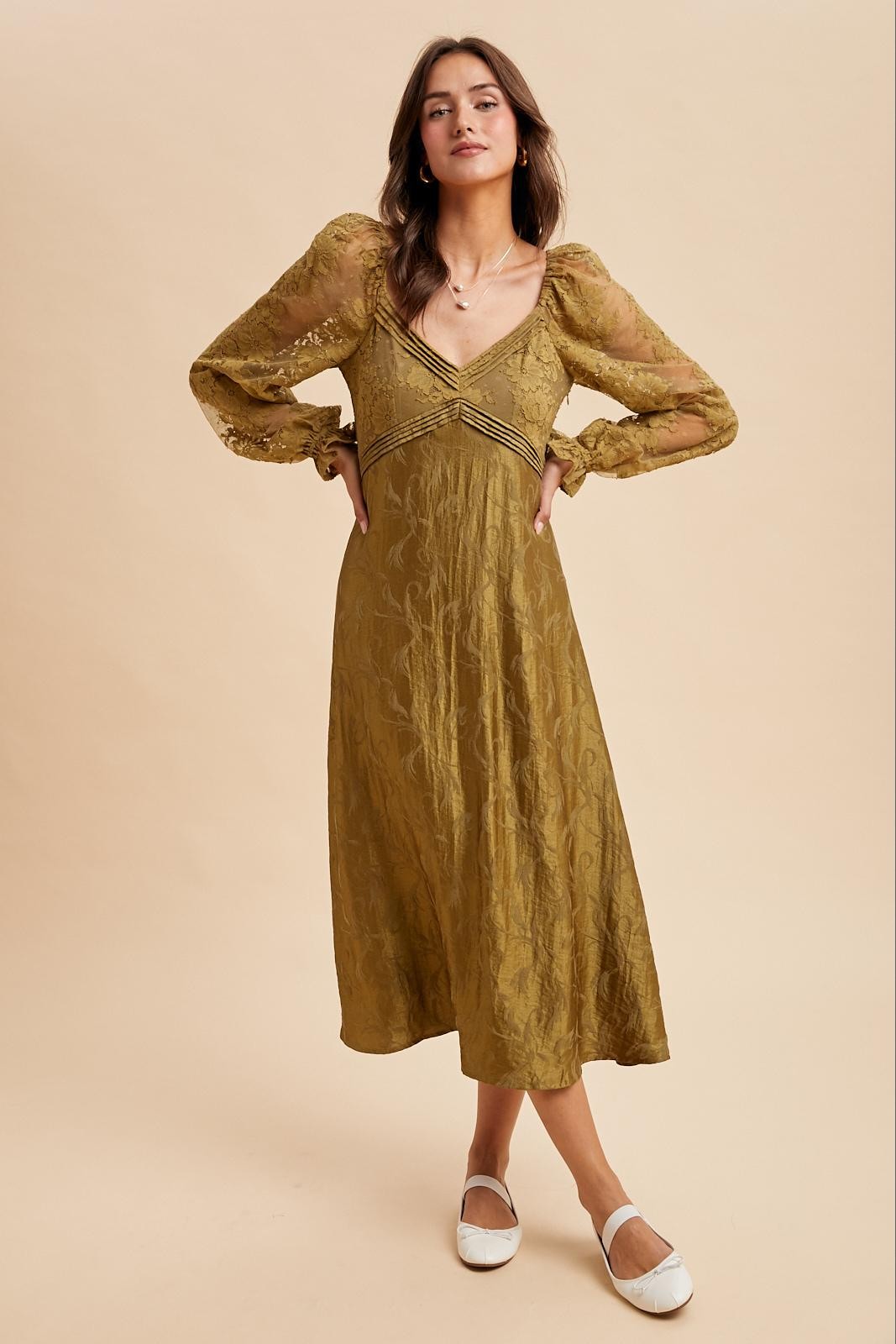 woman wearing a long olive green dress with sheer puff sleeves and lace detailing, standing on a light background
