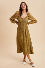 woman wearing a long olive green dress with sheer puff sleeves and lace detailing, standing on a light background