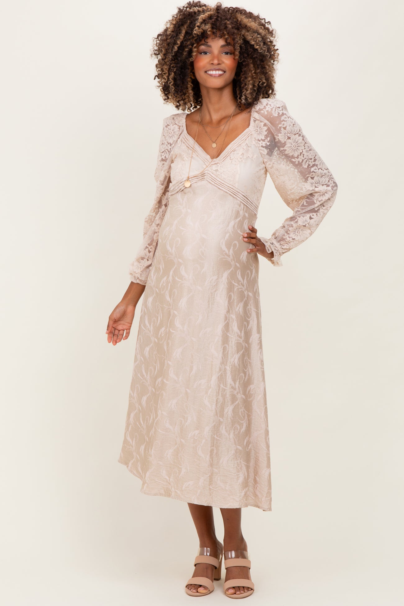 woman wearing a light beige maternity dress with lace long sleeves and a flowy skirt, showcasing her baby bump