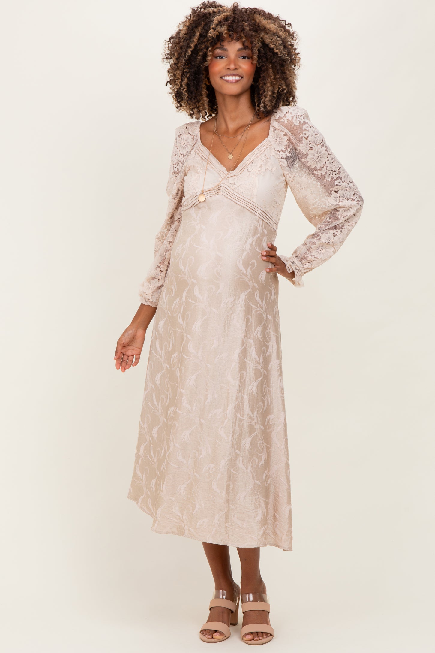 woman wearing a light beige maternity dress with lace long sleeves and a flowy skirt, showcasing her baby bump