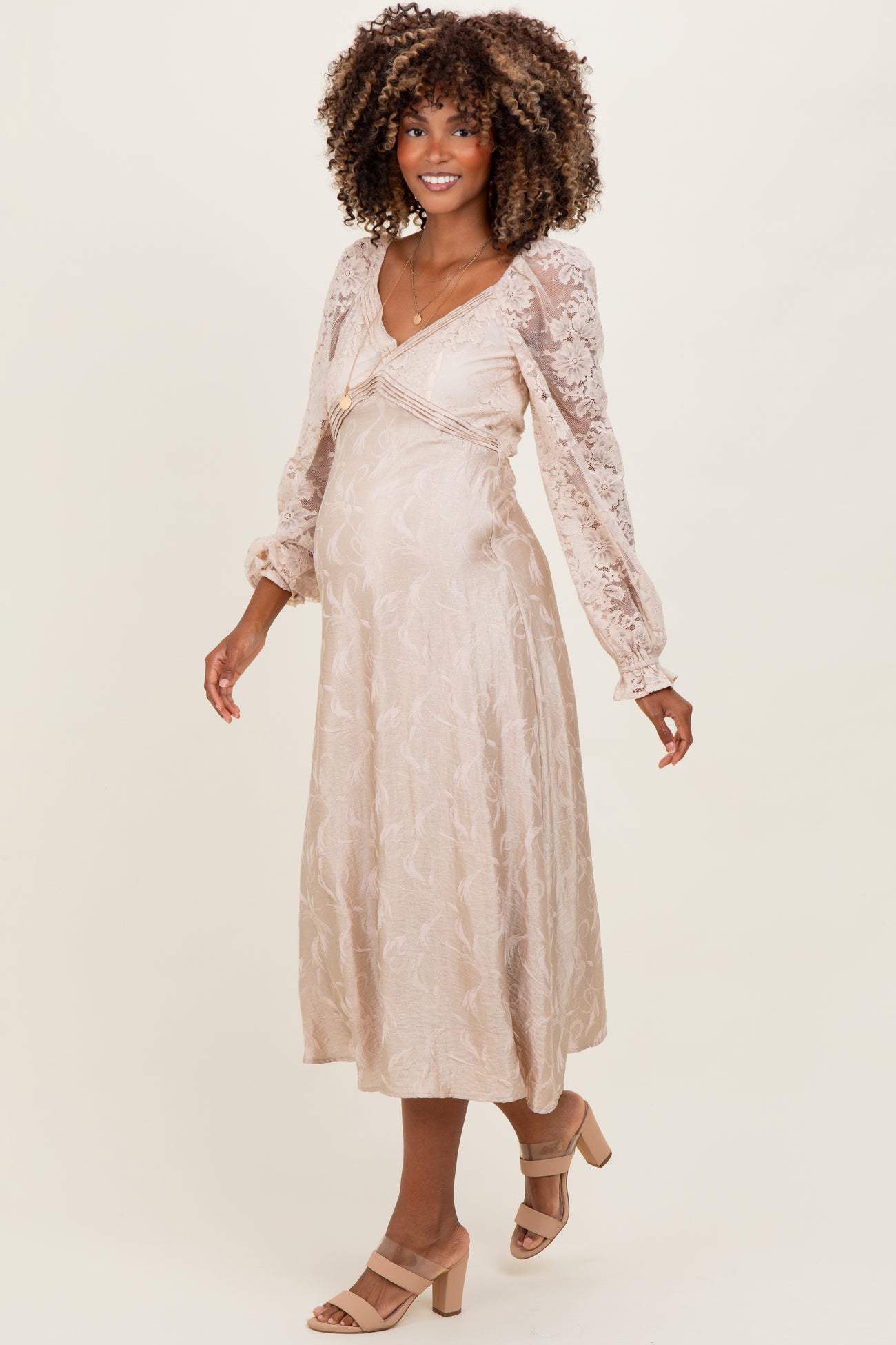 woman wearing a light beige maternity dress with long sleeves and floral lace detail, showcasing her baby bump