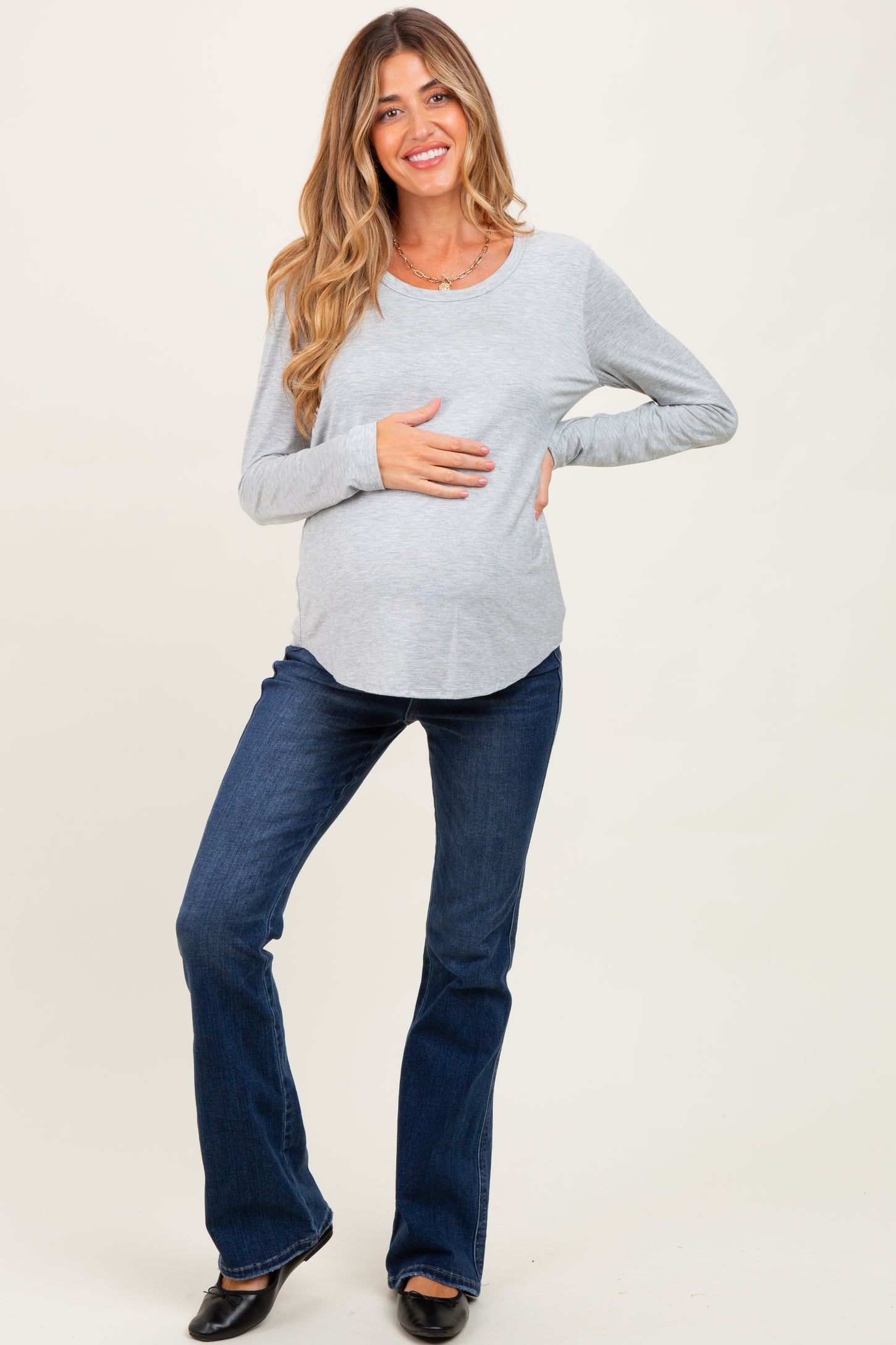 woman wearing a gray long sleeve maternity top and flared jeans, holding her baby bump