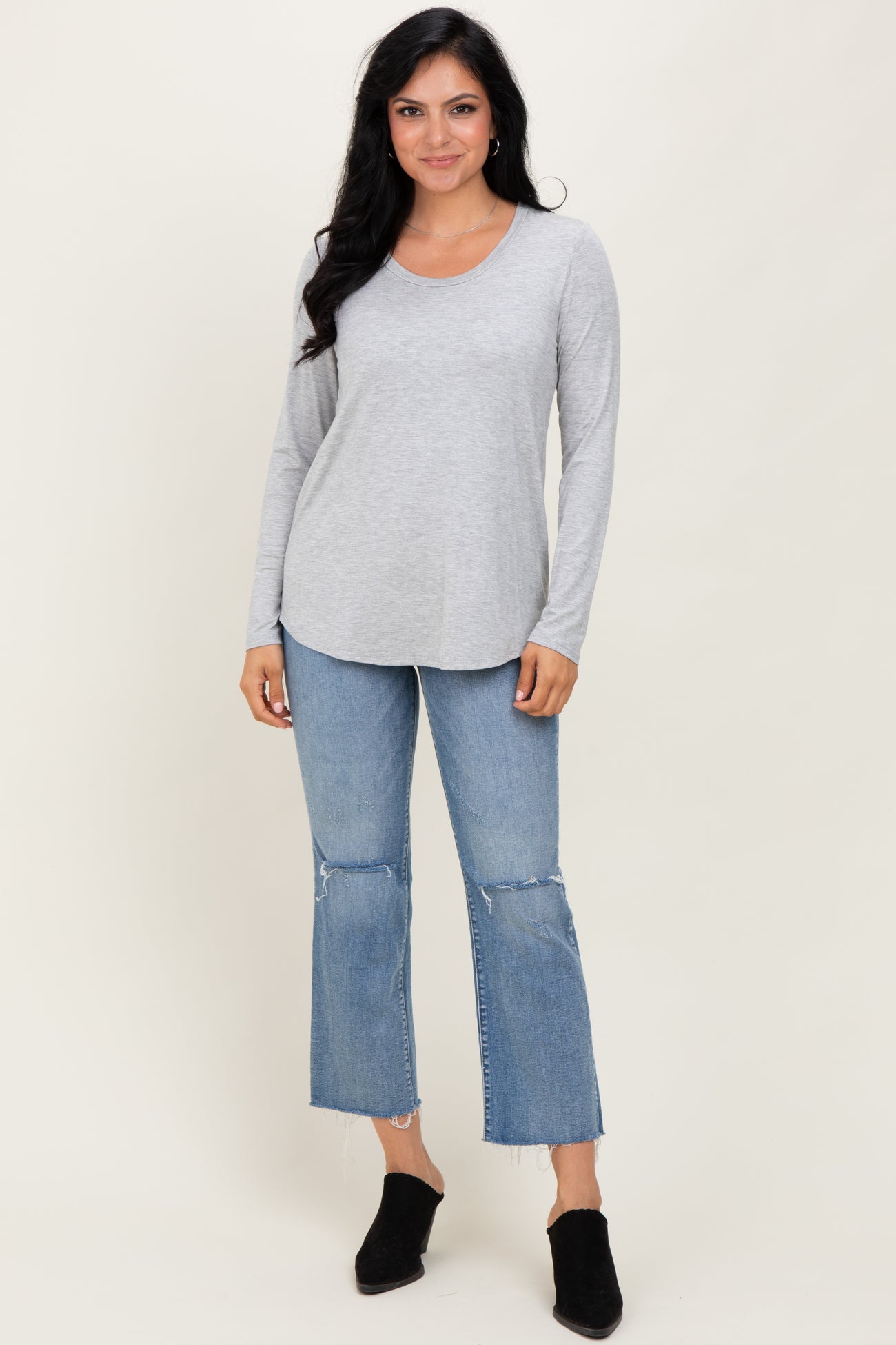 woman wearing a light gray long sleeve top with a curved hem paired with distressed blue jeans and black ankle boots