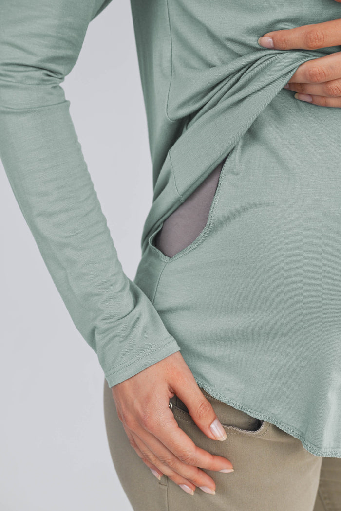 woman wearing a soft green long sleeve maternity top with side pockets, gently holding her baby bump