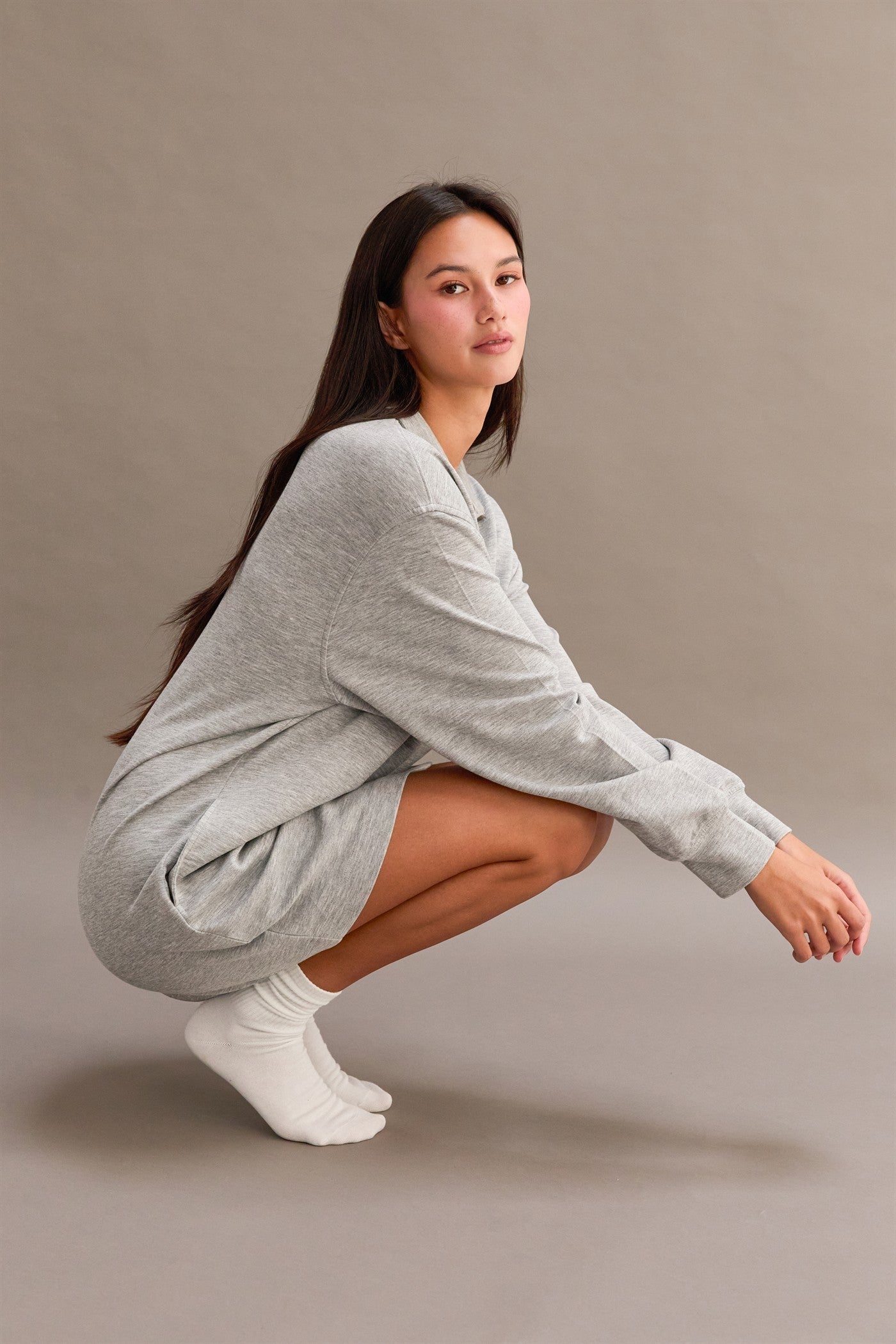 woman wearing a gray long sleeve oversized sweatshirt dress, sitting in a squatting position with white socks
