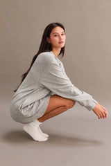 woman wearing a gray long sleeve oversized sweatshirt dress, sitting in a squatting position with white socks
