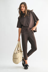 woman wearing a brown loose-fitting t-shirt and matching tapered leggings, holding a beige gym bag