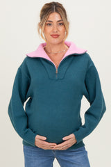 woman wearing a teal maternity pullover with a pink collar and holding her baby bump