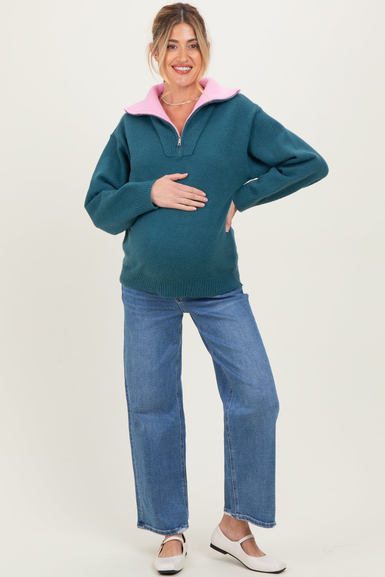 woman wearing a teal maternity pullover with a pink collar and holding her baby bump, paired with jeans