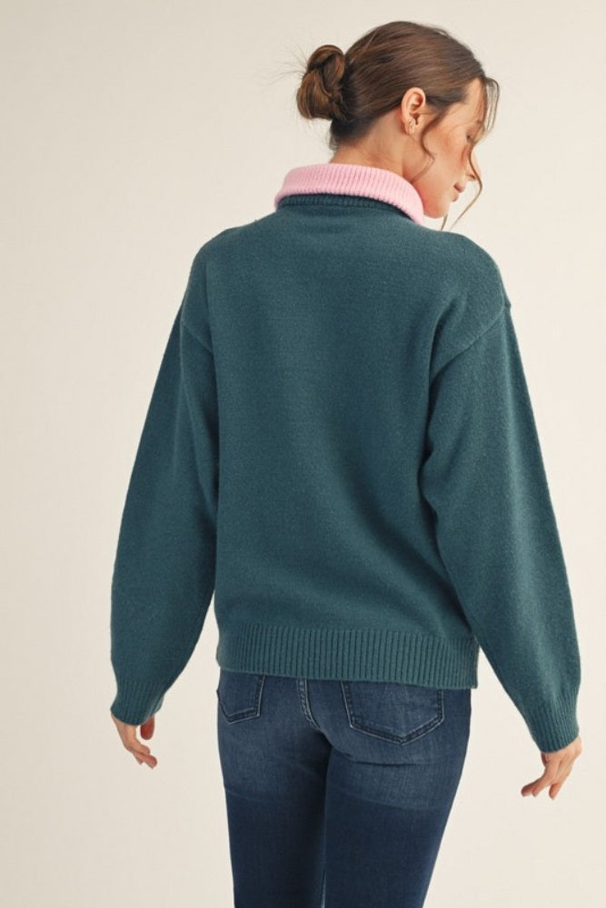 woman wearing a teal oversized sweater with a pink collar and back turned to the camera, paired with dark jeans