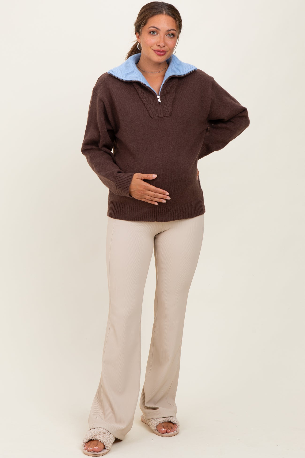 woman wearing a brown maternity pullover with a blue collar and beige maternity pants, holding her baby bump