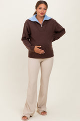 woman wearing a brown maternity pullover with a blue collar and beige maternity pants, holding her baby bump