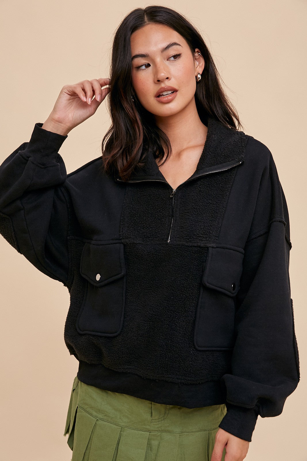 woman wearing a black zip-up pullover with fleece patch pockets and a relaxed fit, paired with a green pleated skirt