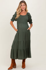 woman wearing a dark green maternity maxi dress with puff sleeves and tiered skirt, holding her baby bump