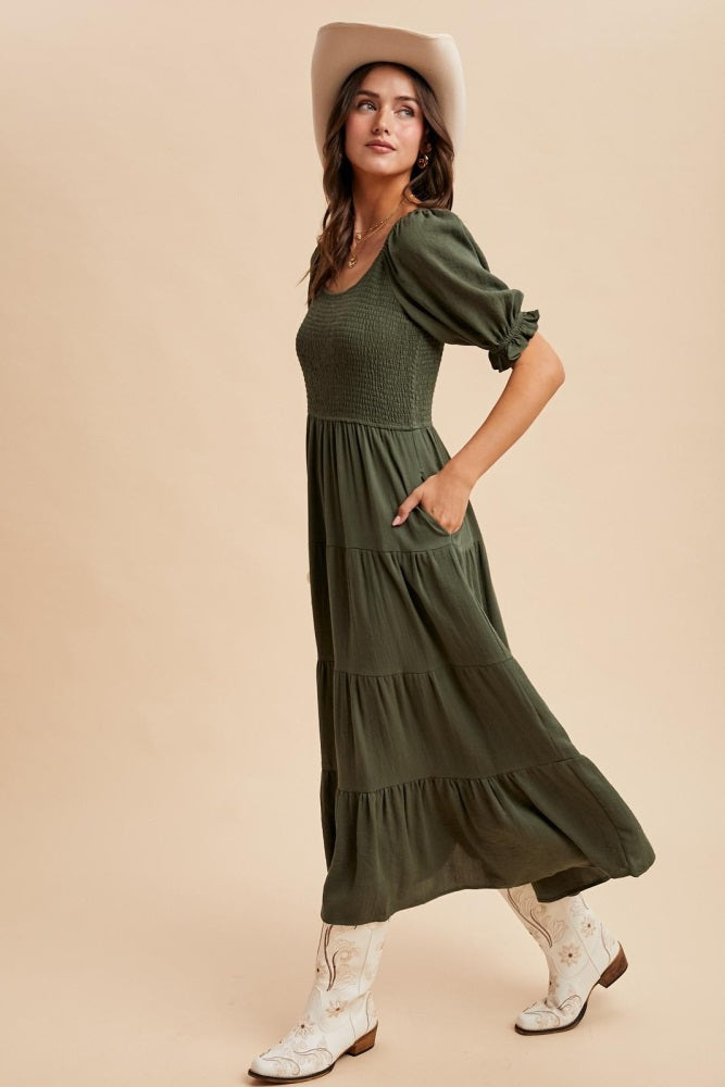 woman wearing an olive green tiered maxi dress with puff sleeves and side pockets, paired with white cowboy boots and a hat