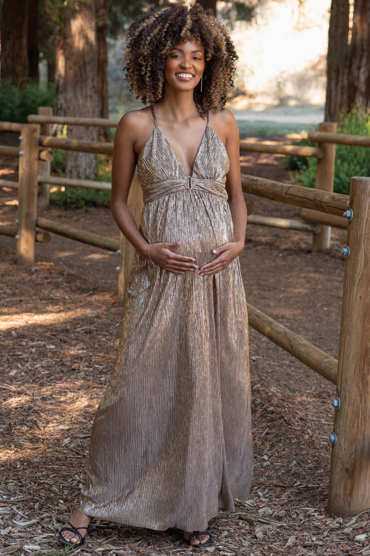 woman wearing a shimmery bronze maternity maxi dress and holding her baby bump in an outdoor setting