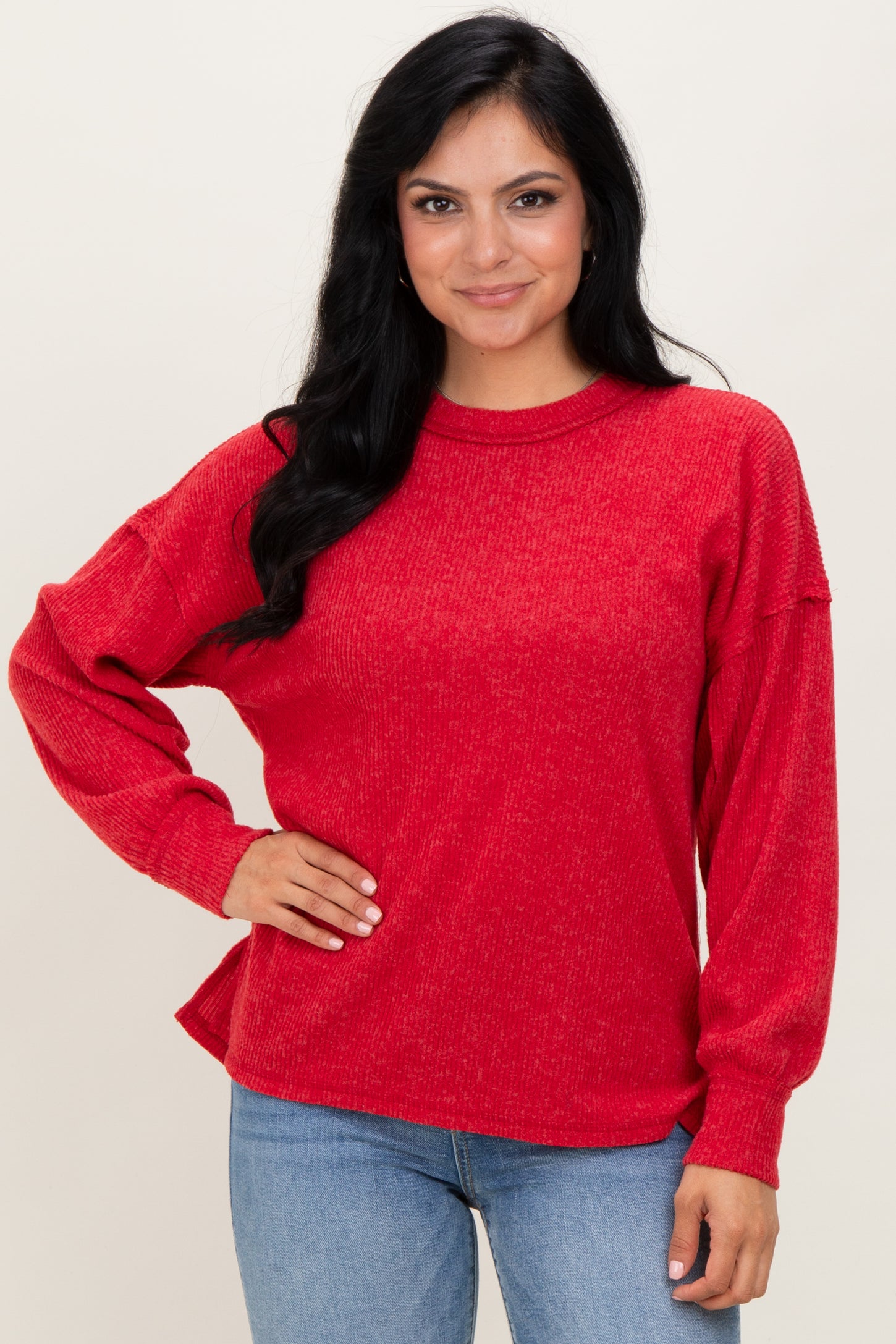 woman wearing a red relaxed fit long sleeve sweater with a textured fabric and side slits, paired with blue jeans