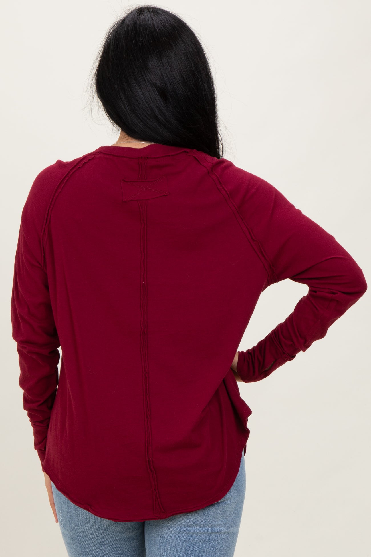 woman wearing a burgundy long sleeve top with a rounded hem and stitch detailing, view from the back