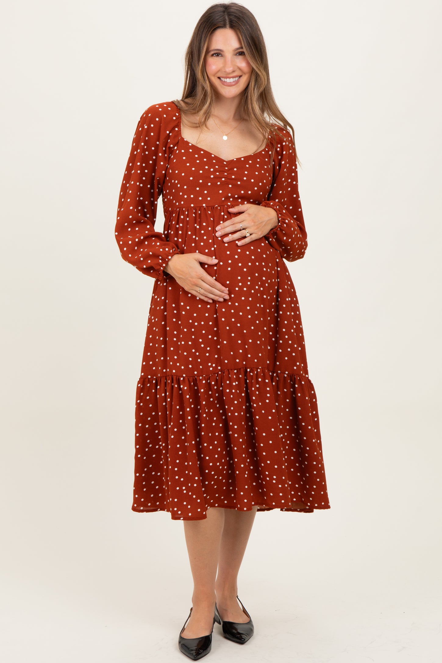 woman wearing a rust polka dot maternity dress with a tiered hem and long sleeves, holding her baby bump