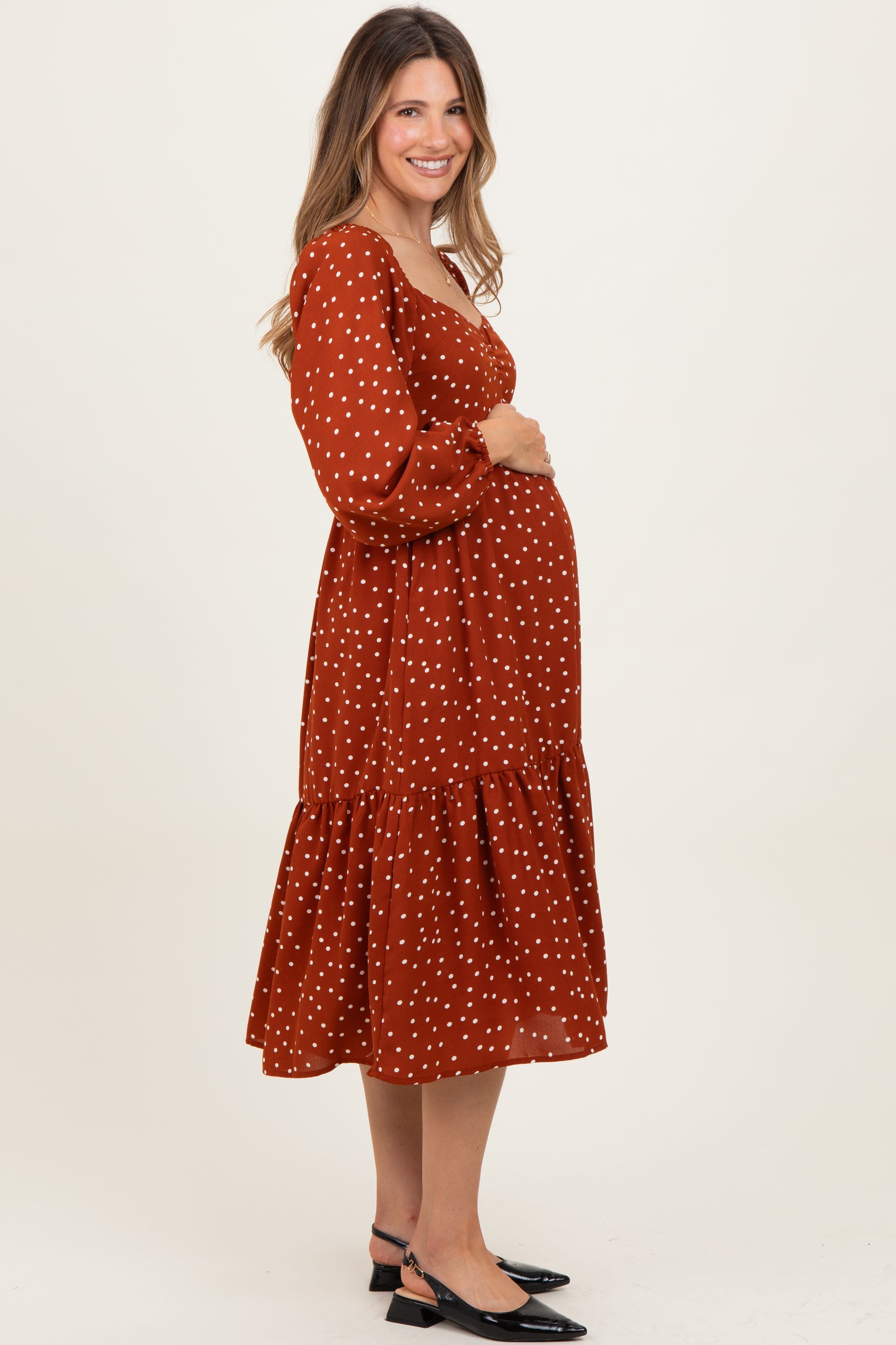 woman wearing a rust polka dot maternity dress with puff sleeves and tiered skirt, cradling her baby bump