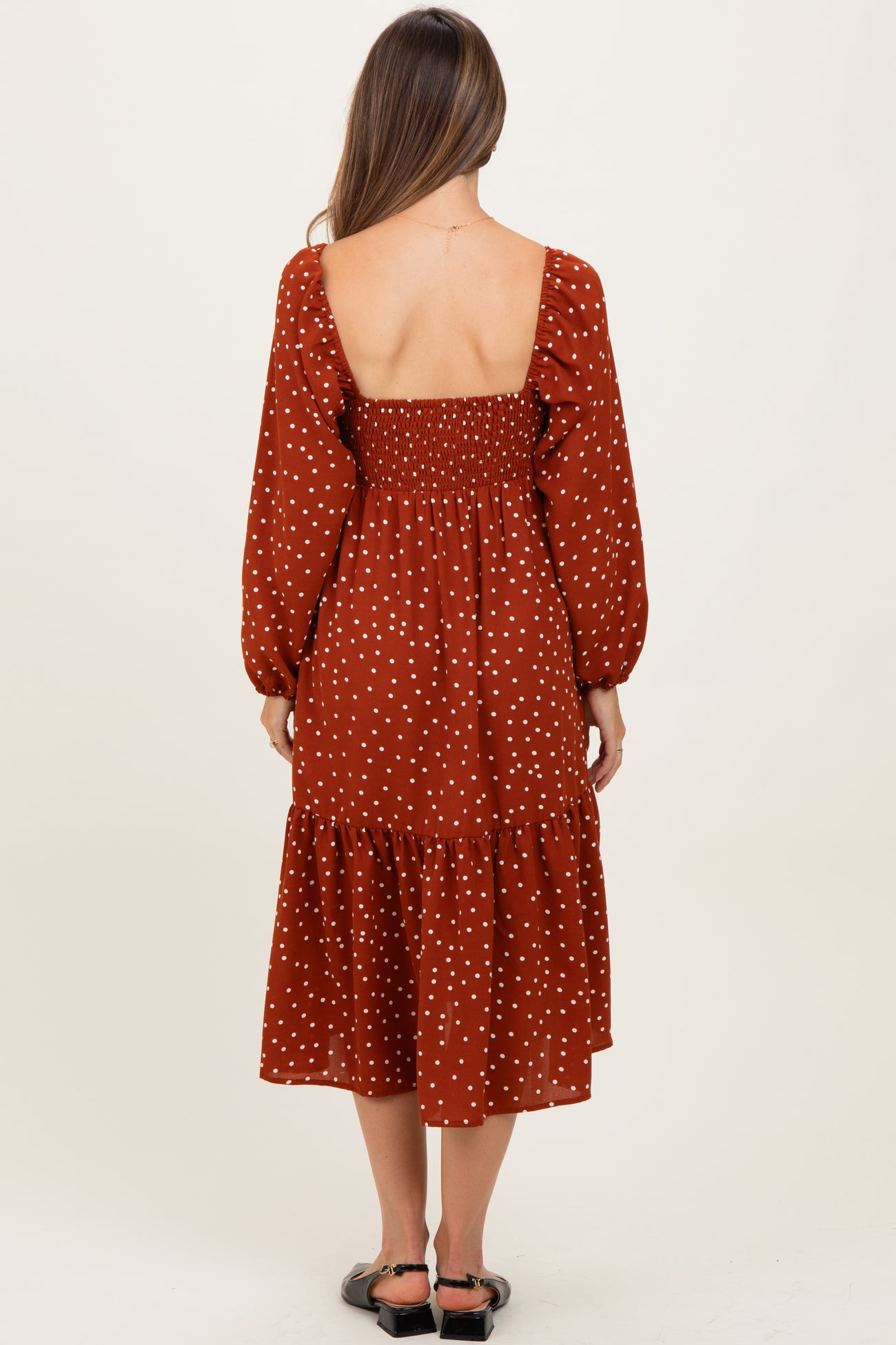 woman wearing a rust-colored maternity dress with polka dots and a puff sleeve, showing a smocked back and tiered skirt