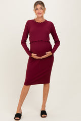 woman wearing a burgundy ribbed maternity dress with long sleeves and a scalloped hem, holding her baby bump