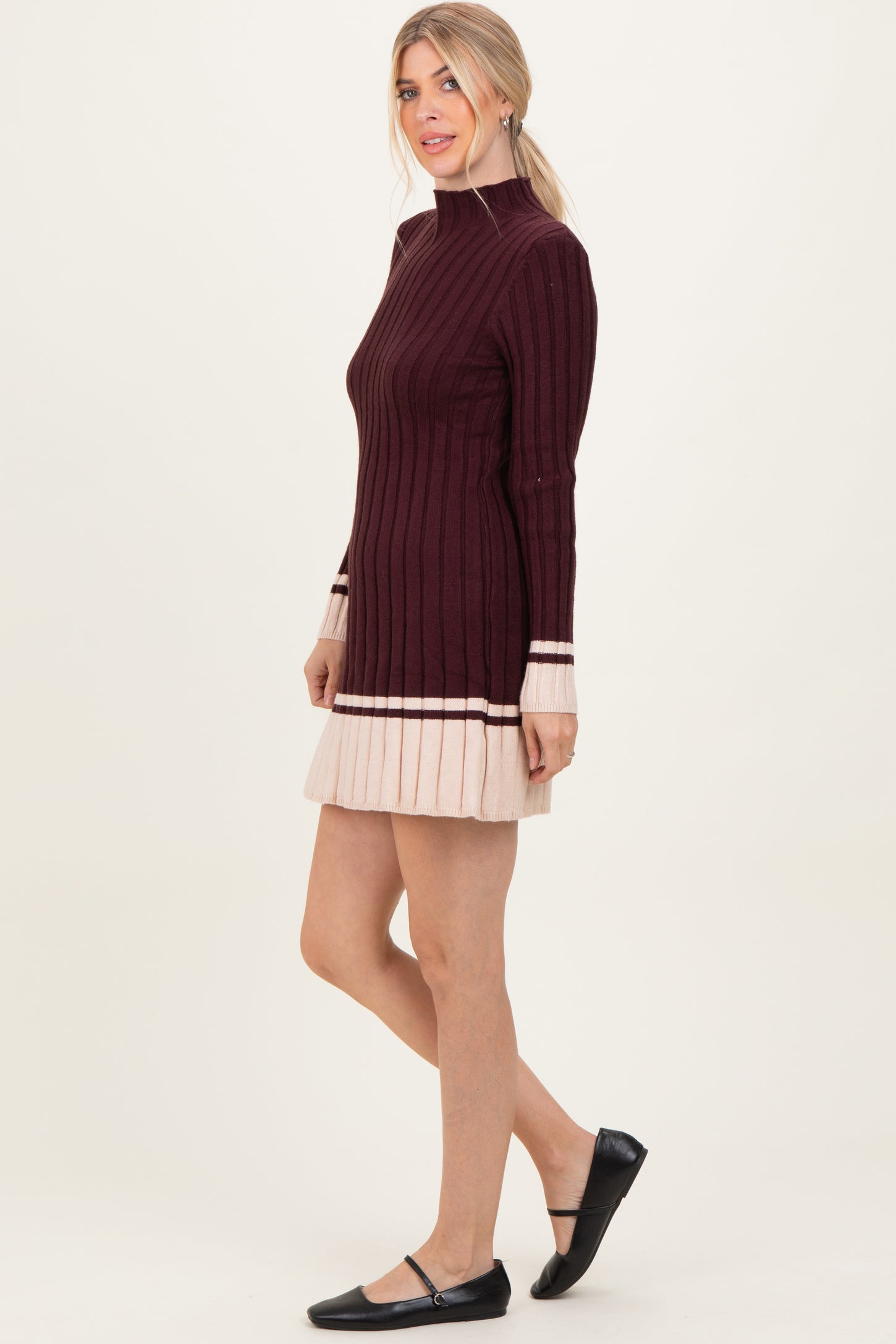 woman wearing a burgundy ribbed long sleeve sweater dress with a pleated beige hem and black flats