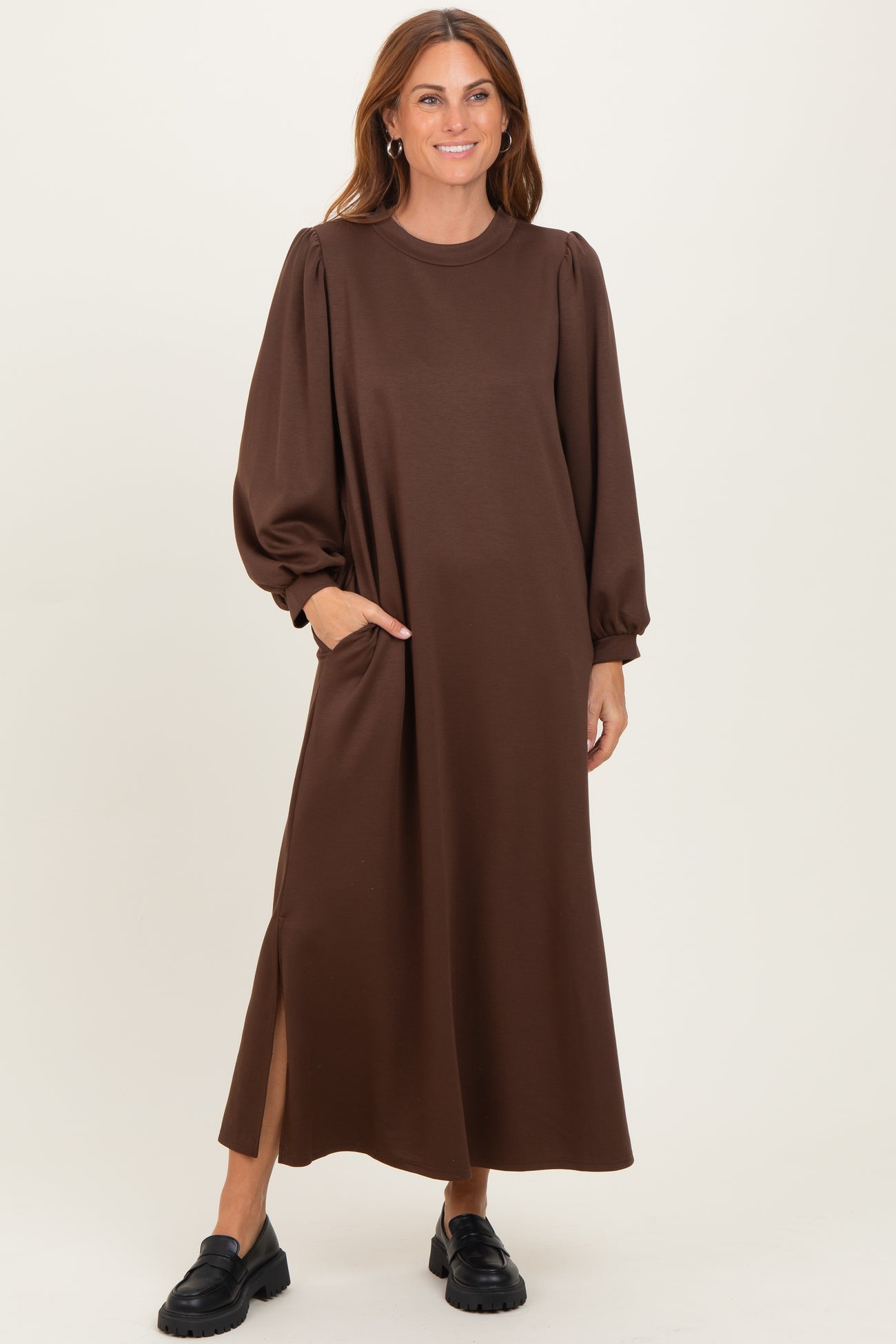woman wearing a brown long sleeve maxi dress with puff sleeves and side pockets, standing and smiling